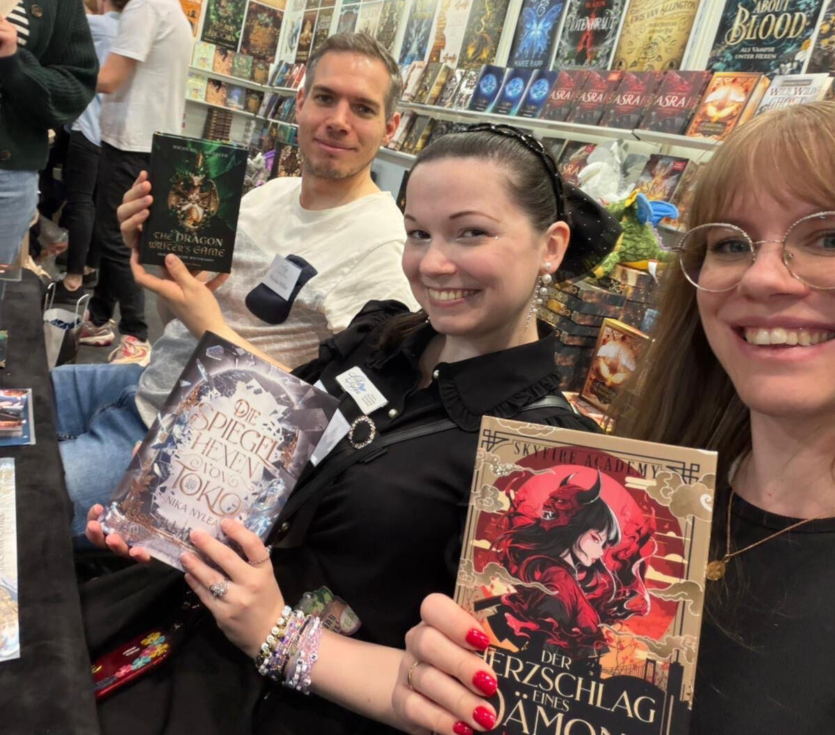 Nicolas Mueller, Nika Nylea und Jenny Pieper sitzen am Stand des Drachenmond Verlags auf der Leipziger Buchmesse. Sie haben eine Signierstunde und halten ihre Bücher in den Händen.