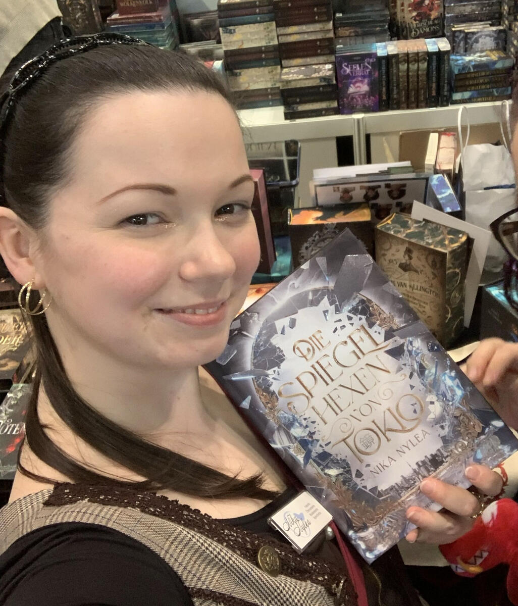Nika hält ihr Buch "Die Spiegelhexen von Tokio" in der Hand. Hinter ihr sieht man den Stand des Drachenmond Verlags auf der Leipziger Buchmesse.