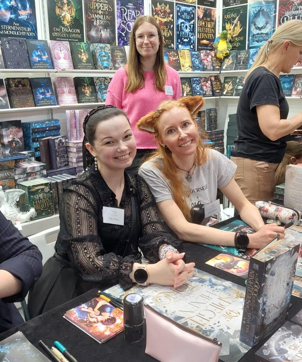 Nika Nylea und Jenny Rubus sitzen beim Stand des Drachenmond Verlags auf der Leipziger Buchmesse. Sie haben eine gemeinsame Signierstunde.