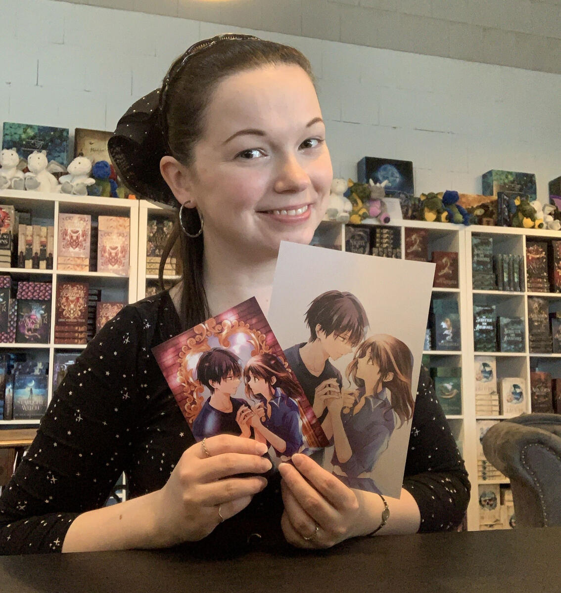 Nika hält die Charakterkarte sowie das Page Overlay von "Die Spiegelhexen von Tokio", gezeichnet von Meitei Miu, in der Hand. Sie sitzt beim Drachenmond Verlag.