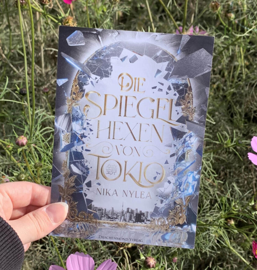 Das Cover von Nika Nylea's "Die Spiegelhexen von Tokio" auf einer Postkarte vor einem Beet